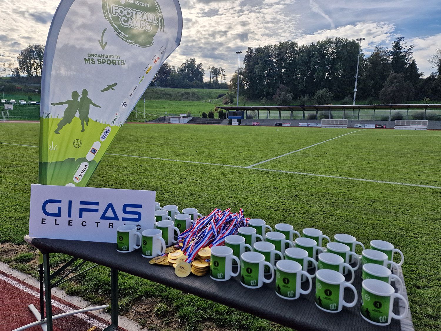 Pokale, Medaillen und Tassen für eine Siegerehrung auf dem Sportplatz mit sichtbarer Eventbeleuchtung im Hintergrund.